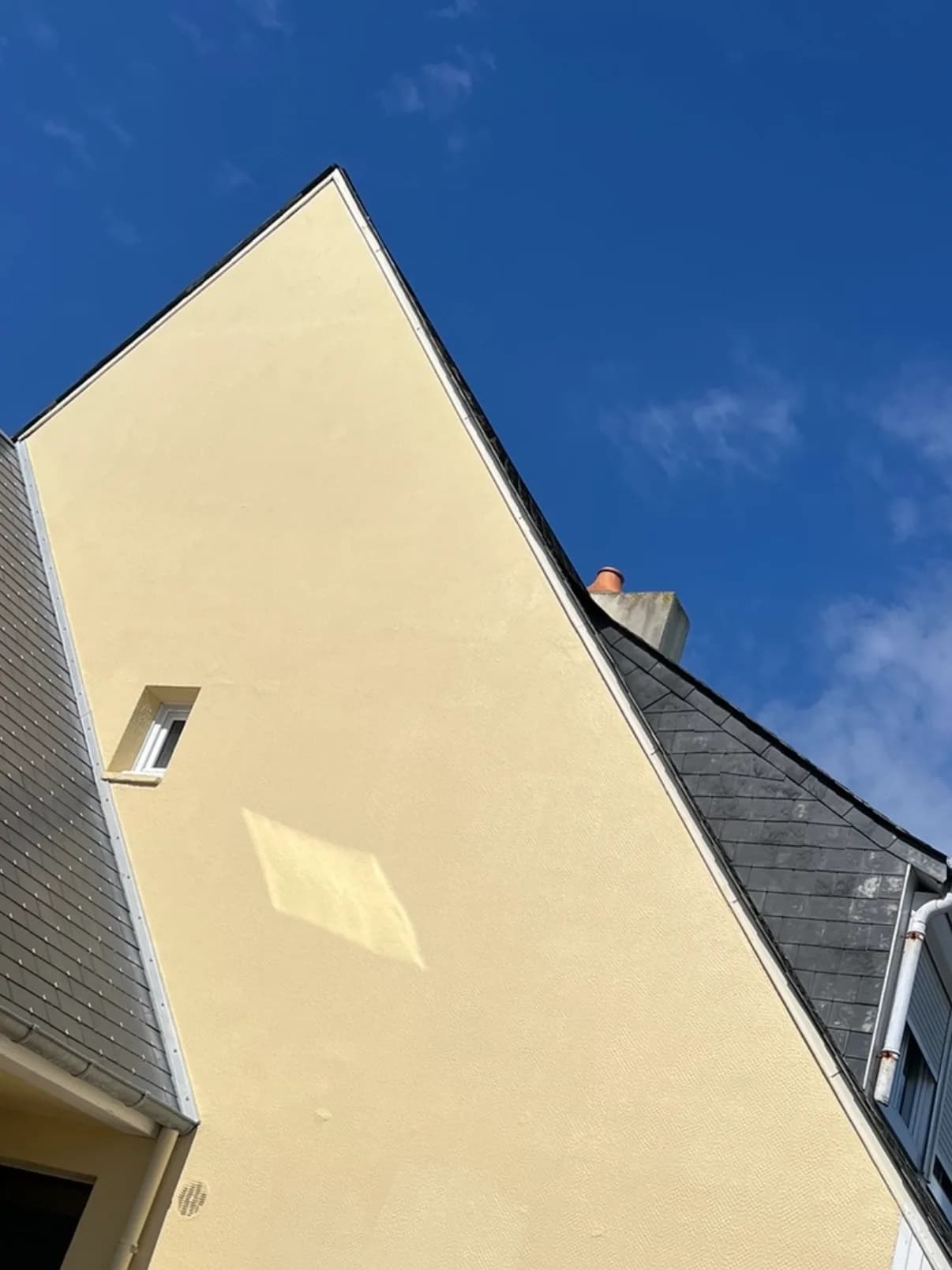 Le même pignon repeint en jaune paille sous un ciel bleu, un peintre passe le rouleau sur perche au pied du mur